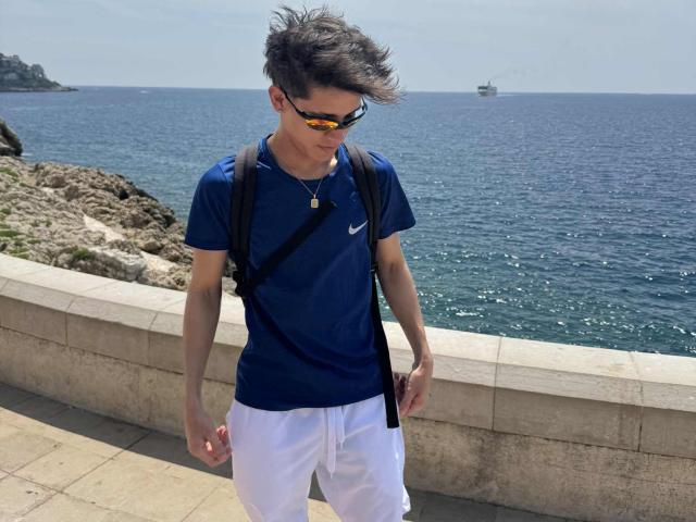 WillStarr wearing a blue Nike t-shirt, white shorts, black backpack, and orange-tinted sunglasses, with short dark hair, standing on a stone pathway by the blue sea.