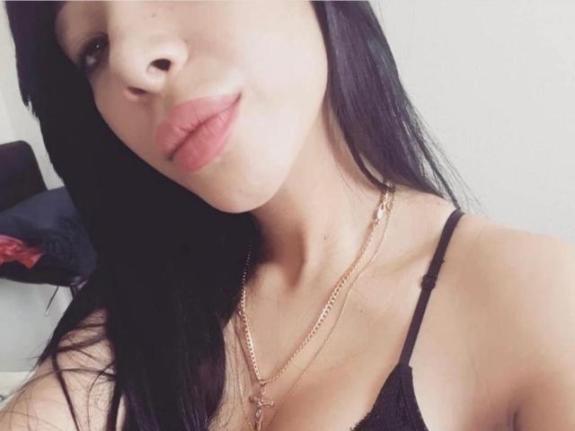 MistressKattia with long black hair, pink lips, wearing a black spaghetti strap top, gold necklace, and showing her small tits. Her face is slightly angled to the left.