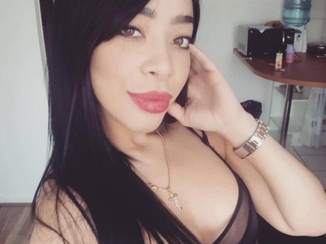 MistressKattia with long black hair, red lipstick, and a gold necklace, showing off her tits in a sheer black top, touching her face with a gold watch.