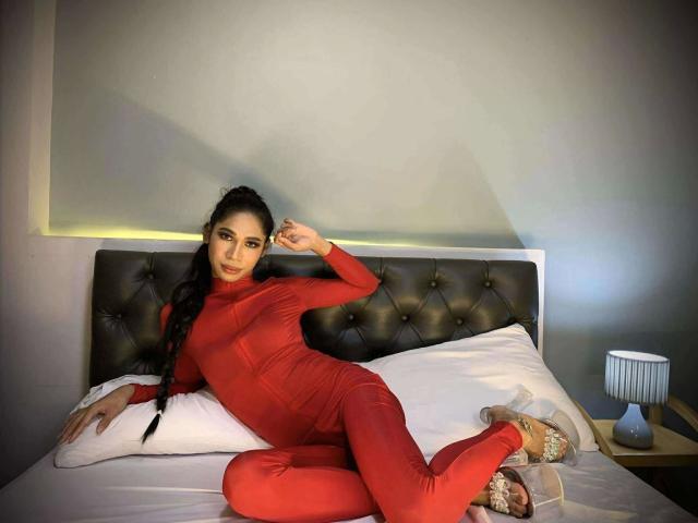 JulietMonte - Sensuelle dans une combinaison rouge moulante à manches longues, allongée sur un lit avec des oreillers blancs, cheveux noirs tressés, talons hauts couleur chair, une main dans les cheveux, sur une tête de lit capitonnée..