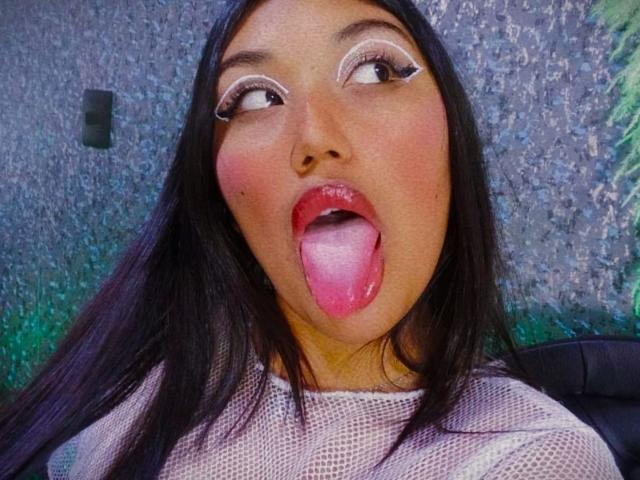 SharaRous, com cabelos pretos longos, maquiagem e olhos grandes, língua de fora, língua rosa, vestindo uma blusa branca de malha, olhando para cima, pele morena, fofa, sexy, close-up do rosto..