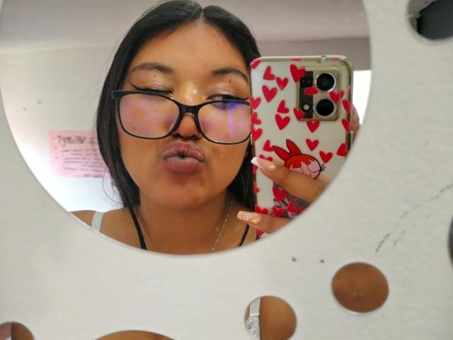 SharaRous, con cabello oscuro, gafas y labios carnosos, sostiene un teléfono con una funda roja con estampado de corazones, y su reflejo se ve en un espejo blanco con forma recortada. Sus uñas son largas y brillantes..