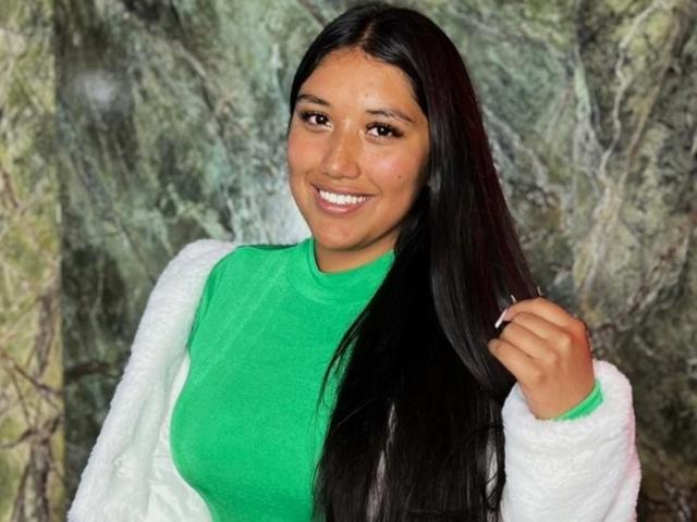 SharaRous with long black hair, green turtleneck, white fluffy jacket, smiling, holding hair, brown eyes, medium brown skin, cute, attractive, sexy.