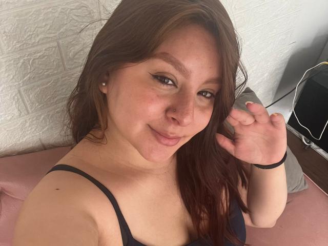 EmaJames with brown hair, smiling, nose ring, black strap dress, raising right hand, showing off small tits and cute face, pink pillow, white textured wall, and black speaker.