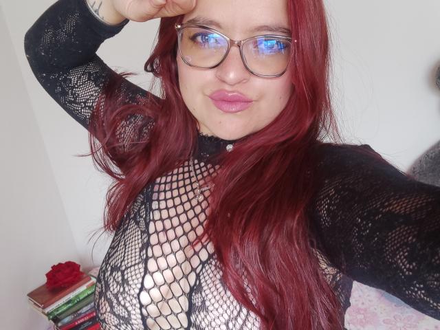 ElizabethFire with red hair, glasses, and pink lips, wearing a black fishnet top that reveals her tits, slightly smiling, raising her right hand to her forehead.