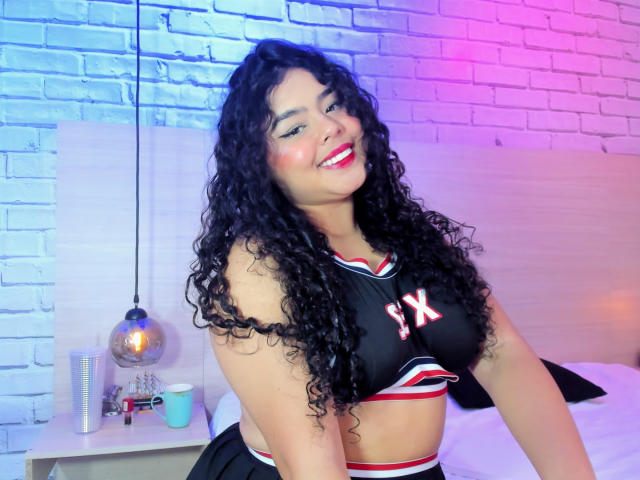 AprielKeith with curly black hair, smiling, wearing a black and red sports bra with "1X" and matching skirt, showing her tits, in a brightly lit room.