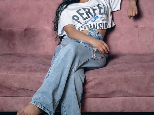 NatallyCooper with long black hair, white t-shirt, and ripped blue jeans, sitting on a pink suede couch, showing her ass and slightly visible tan lines.