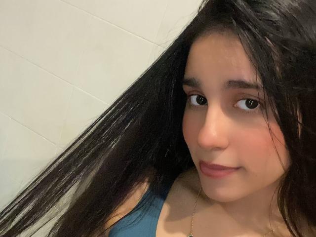 TiffannyRose has long black hair, brown eyes, and a slight smile. She's wearing a blue top, and her small tits are partially visible. She's looking at the camera.