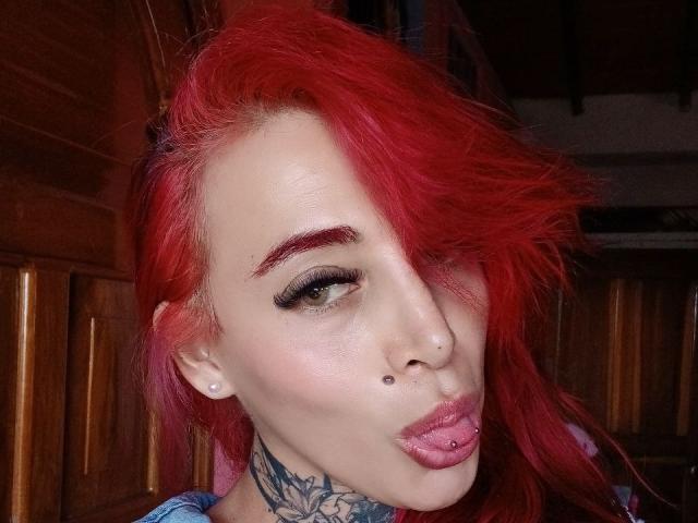 YeiSela with bright red hair, green eyes, and pink lip piercings, wearing black eyeliner, nose stud, and lip stud, showing off a tattooed neck. Wooden background.