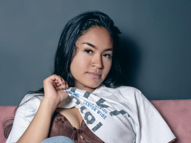 Natally Cooper, met lang zwart haar, bruine huid en een kleine neus, tilt een wit shirt op om een bruine kanten bh te laten zien, waarbij haar borsten zichtbaar zijn, terwijl ze op een roze bank zit..