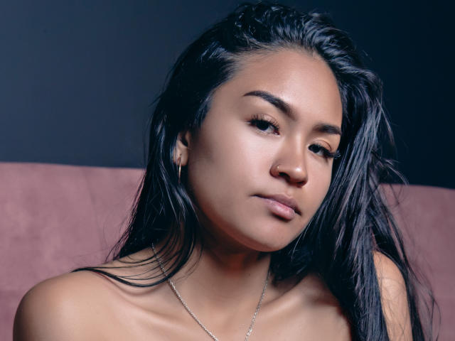 Natally Cooper, con cabello negro largo, piel morena y una nariz pequeña, lleva un collar, tiene labios carnosos y una expresión ligeramente fruncida, mirando hacia un lado, mostrando sus hombros desnudos..