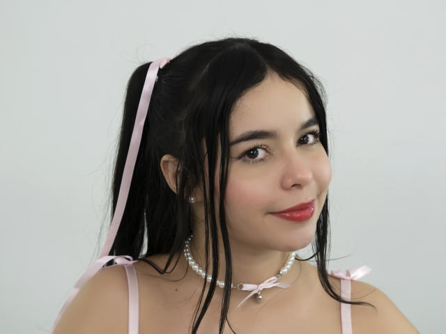 LissyCarter with black hair in a pink ribbon ponytail, wearing a pink strap dress, pearl necklace, and red lipstick, smiling softly. Her tits are small, and she's cute.