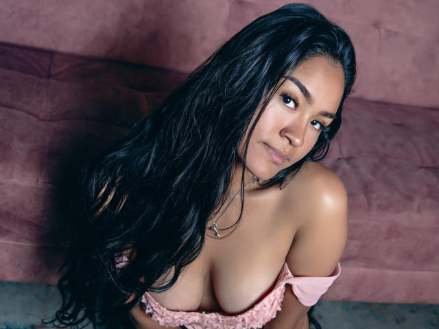Natally Cooper, con lunghi capelli neri, pelle abbronzata e seno prosperoso, indossa una maglietta rosa a spalle scoperte, è seduta su un divano rosa e guarda verso l'alto con un sorriso appena accennato..