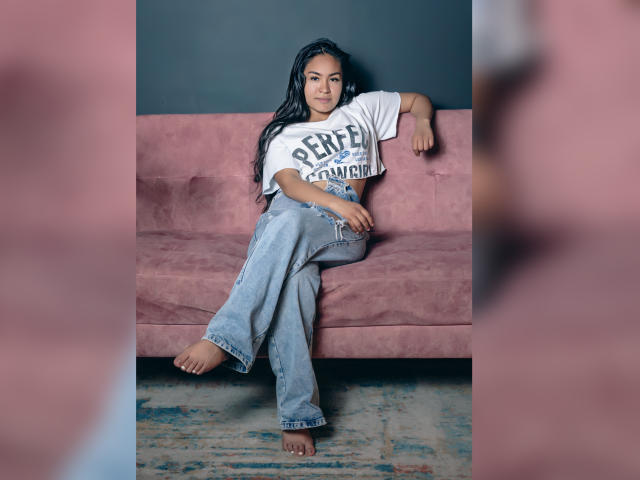 Natally Cooper, con i suoi lunghi capelli neri, è appoggiata a un divano rosa, indossando una maglietta bianca con la scritta "CEFA" e jeans blu larghi, a piedi nudi, con le gambe incrociate, e appare rilassata e sicura di sé..