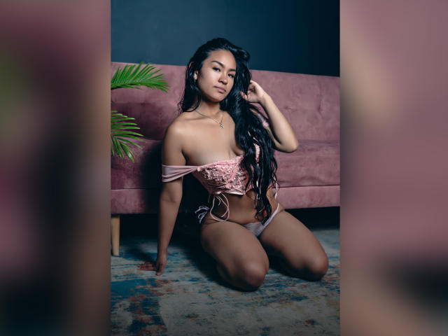 NatallyCooper, con lunghi capelli neri, inginocchiata su un tappeto, indossa una maglietta rosa a spalle scoperte e biancheria intima coordinata, mostrando i suoi piccoli seni e la sua graziosa vulva, in una stanza rosa..