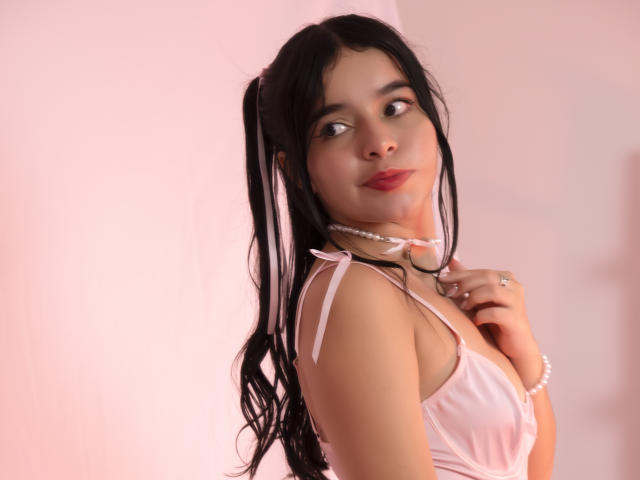 LissyCarter with long black pigtails, pink satin dress, pearl choker, and red lipstick, looking over her shoulder, holding her collar with a ring. Soft pink lighting.