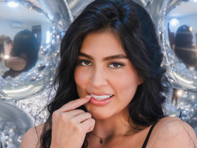 SamanthaEva69 with long black hair, brown eyes, and a cute smile, biting her finger, showing white teeth. She's wearing a black top, with a silver balloon backdrop.