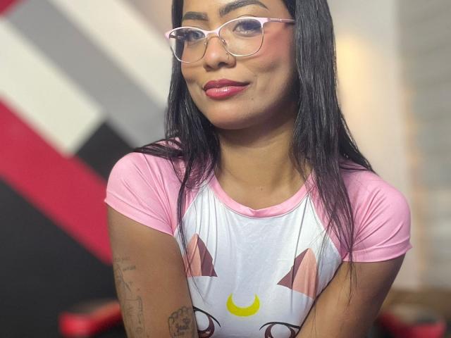 ChloeBrownOne with pink glasses, pink-pink- pink rag tee, and cat face, showing her tits and tattoos, smiling with pink lipstick, and long black hair.