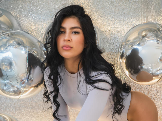 SamanthaEva69 with long black wavy hair, wearing a white long-sleeve bodysuit, and silver balloons reflecting her face. Her ass is prominent, looking over her shoulder.