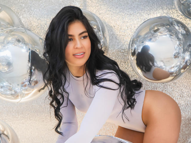 SamanthaEva69 with long black hair, white tight bodysuit, no pants, shiny silver balloons, and a sexy smile, kneeling, showing her ass, with reflective surfaces.