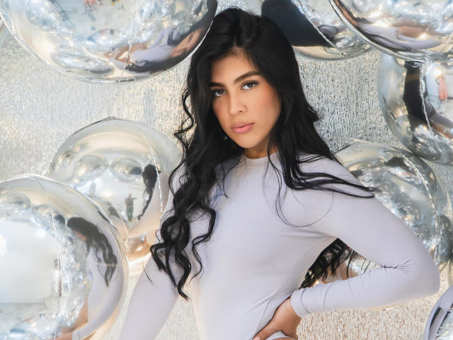 SamanthaEva69 with long black wavy hair, wearing a tight white long-sleeve top, standing in front of shiny silver balloons, looking at the camera with a slight smile.