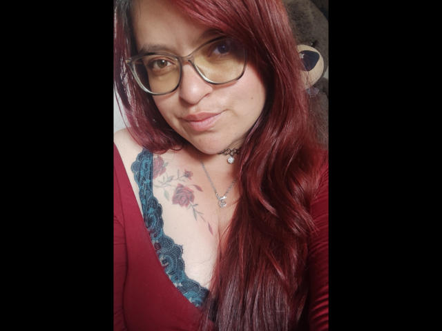 Elizabeth, mit roten Haaren, Brille und einem roten Oberteil, das ihre Brüste zeigt, hat ein Rosentattoo auf ihrer Brust und eine kleine Halskette, und sie lächelt leicht. Ein Teddybär ist im Hintergrund zu sehen..