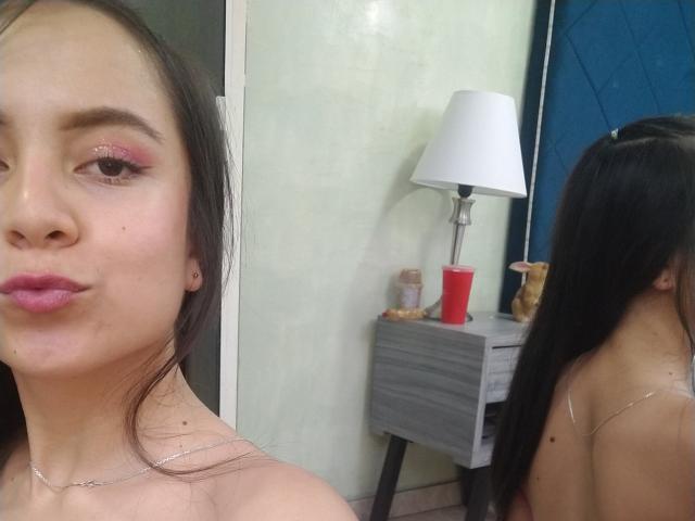 JessyLenon with pink eyeshadow, pink lips, long black hair, mirror reflection showing back, small silver necklace, light skin, subtle makeup, no clothes visible, white table with red cup.