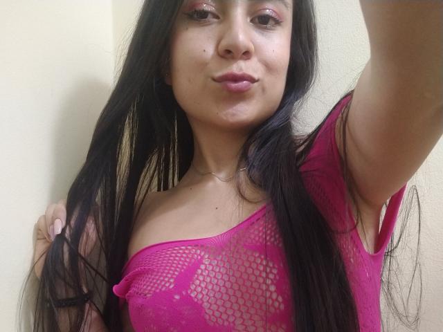JessyLenon with long black hair, pink fishnet top showing her tits, pink eyeshadow, and pouty lips, holding her hair with one arm, against a plain wall.