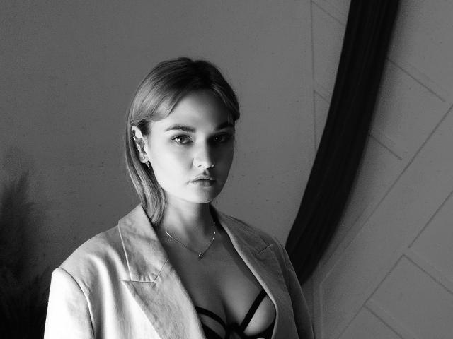 Black-and-white photo of MeriyJewel with light skin, straight blonde hair, small breasts, wearing a strappy black top and open shirt, standing against a wall, looking serious.