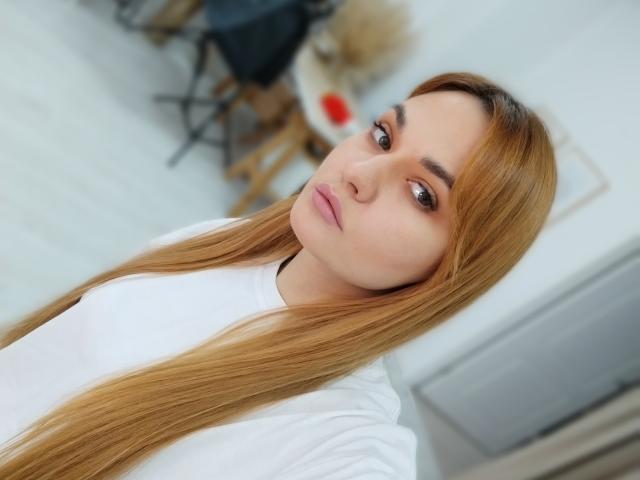 MeriyJewel with long blonde hair, wearing a white shirt, has a neutral expression, brown eyes, and pink lips, standing in a bright room, showing a subtle pout.