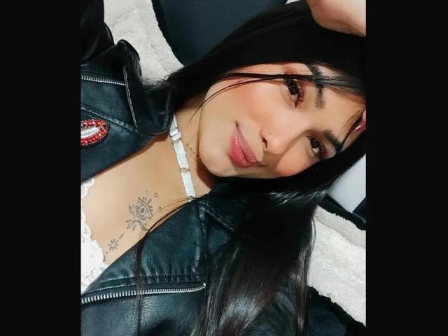 ThianaGray with long black hair, nose ring, white choker, black leather jacket, and rose tattoo on chest, smiling at camera. Boobs visible, wearing lace top.