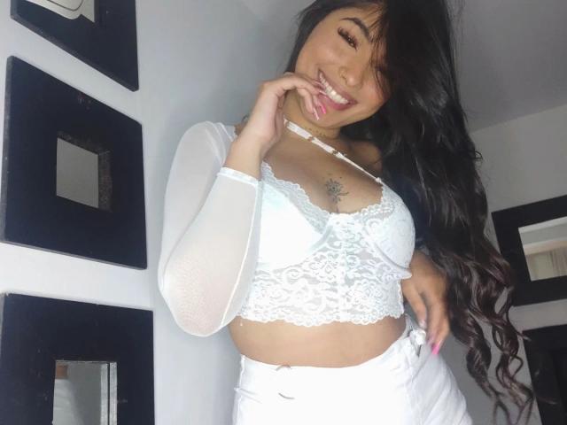 ThianaGray with long black hair, white lace bra, and sheer sleeves, smiling with tongue out, hand near mouth, showing small tattoo under her tits.
