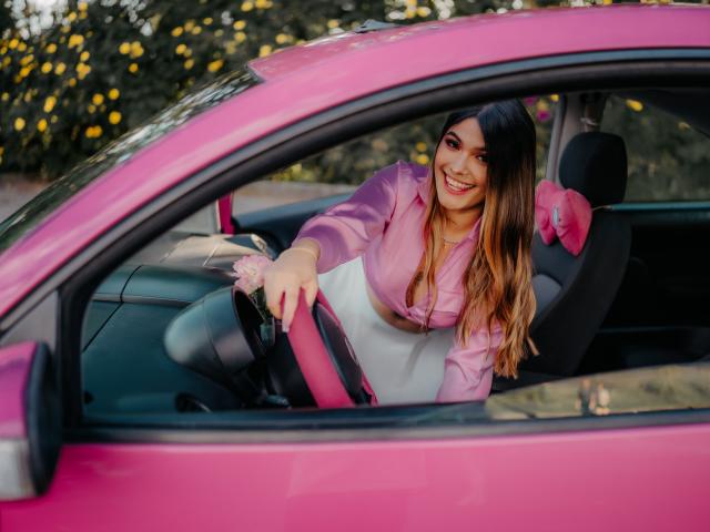 ThianaGray with long brown hair, pink jacket, and white skirt, leaning on pink car steering wheel, smiling, hands on wheel, pink bow on seat, daytime, car window.