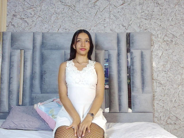 BeautifulIndie with long black hair, wearing a white lace dress, sitting on a bed, legs in fishnet stockings, hands on thighs, smiling, gray headboard, textured wall behind.