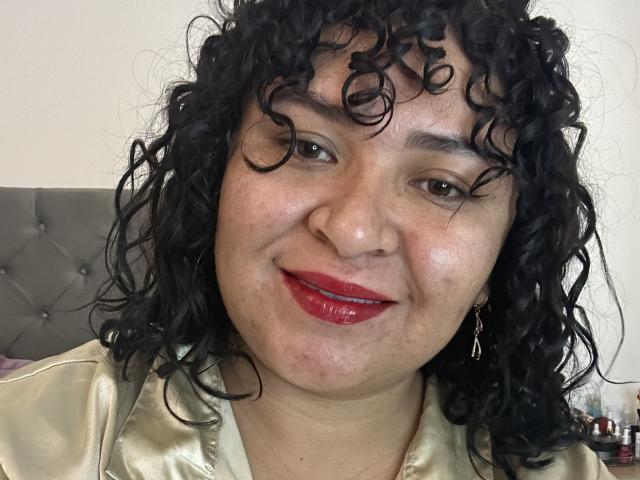 Karla, com cabelos pretos encaracolados, lábios vermelhos e brilhantes, e uma camisa bege brilhante, sorrindo para a câmera. Seus olhos castanhos e a pele levemente avermelhada são visíveis..
