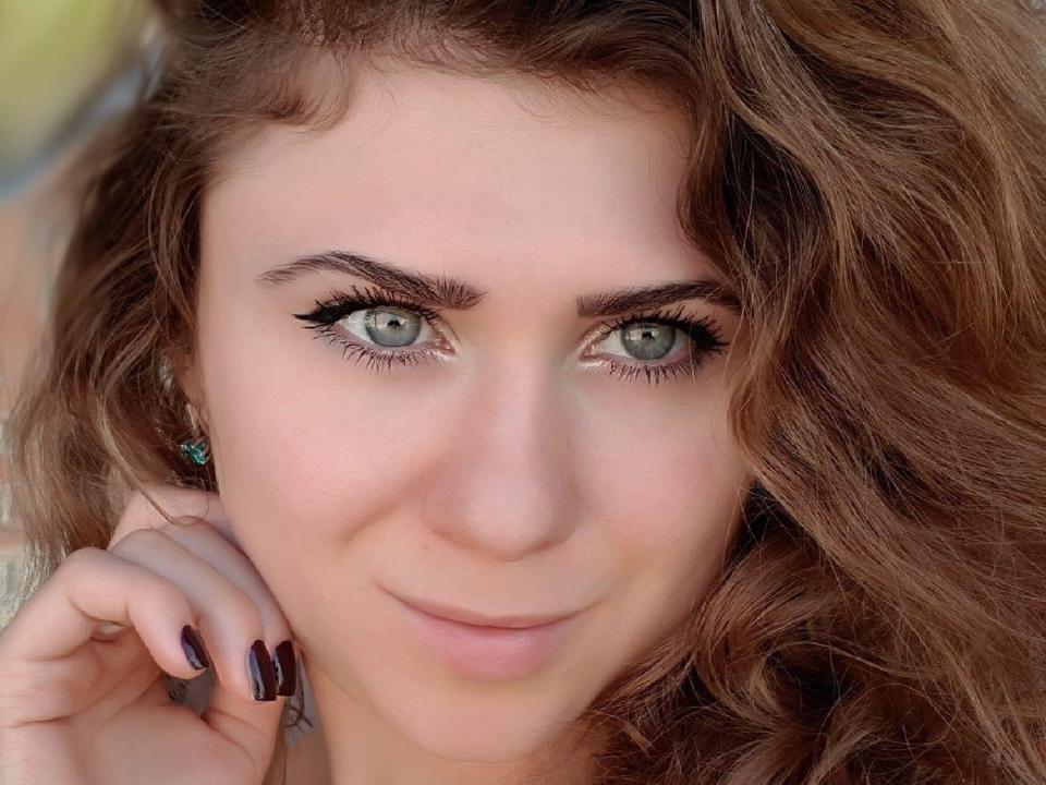 LoriSelma has green eyes, wavy brown hair, and dark eyeliner. She's smiling with a hand near her face, showing dark nail polish and a subtle makeup look.
