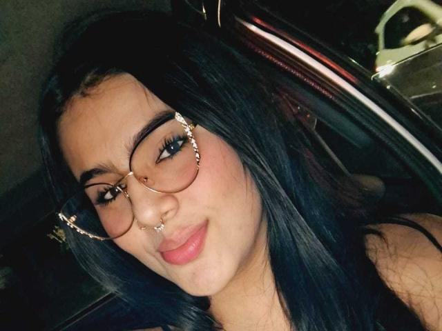 AmberNight, com cabelo escuro, óculos, piercing no nariz e no lábio, sorrindo com lábios rosados, dentro de um carro à noite, vestindo uma blusa preta, mostrando seu rosto adorável..
