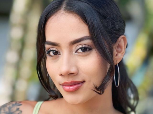LunaGomezz has medium brown skin, black hair in a ponytail, hoop earrings, and red lipstick. She's smiling with subtle makeup and a tattoo on her right shoulder.