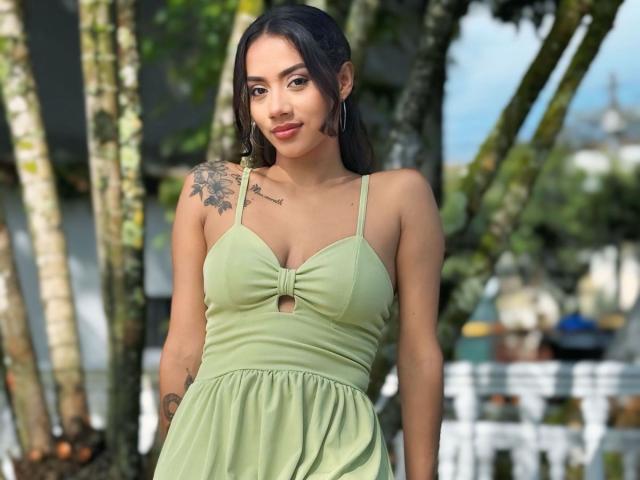 LunaGomezz with small tits, wearing a green dress, has a tattoo on her right shoulder, and large hoop earrings, standing outdoors with trees in the background.