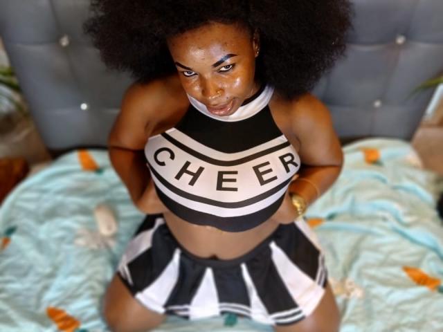 Annykaa with dark skin and big afro, wearing a black and white cheerleader outfit, exposing her big boobs and small black and white skirt, sitting on a bed.