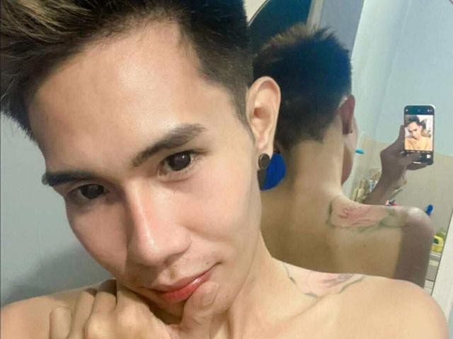ExoticYuki with short brown hair, blue ear studs, and a tattoo on the right shoulder, taking a selfie in a mirror, showing their back and hand on their chin.