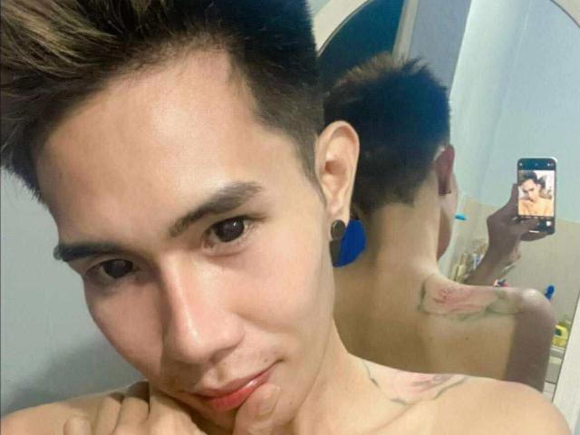 ExoticYuki with short brown hair, blue earrings, and a tattoo on the right shoulder, taking a selfie in a mirror, showing a reflection of their face and back.
