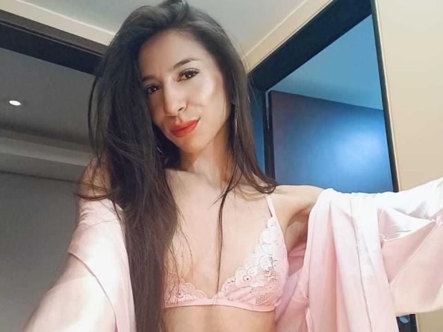NahomyCarther with long brown hair, pink lace bra, and pink robe, showing small tits, smiling at camera, holding robe off shoulders, in a well-lit room.