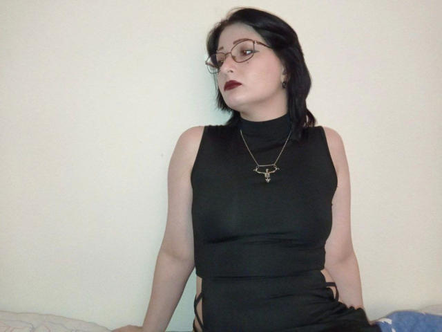 Rachelrocks-hot with dark hair, glasses, red lipstick, black sleeveless turtleneck, necklace, sitting, pale skin, looking up, black dress with side cutouts, exposing her tits.