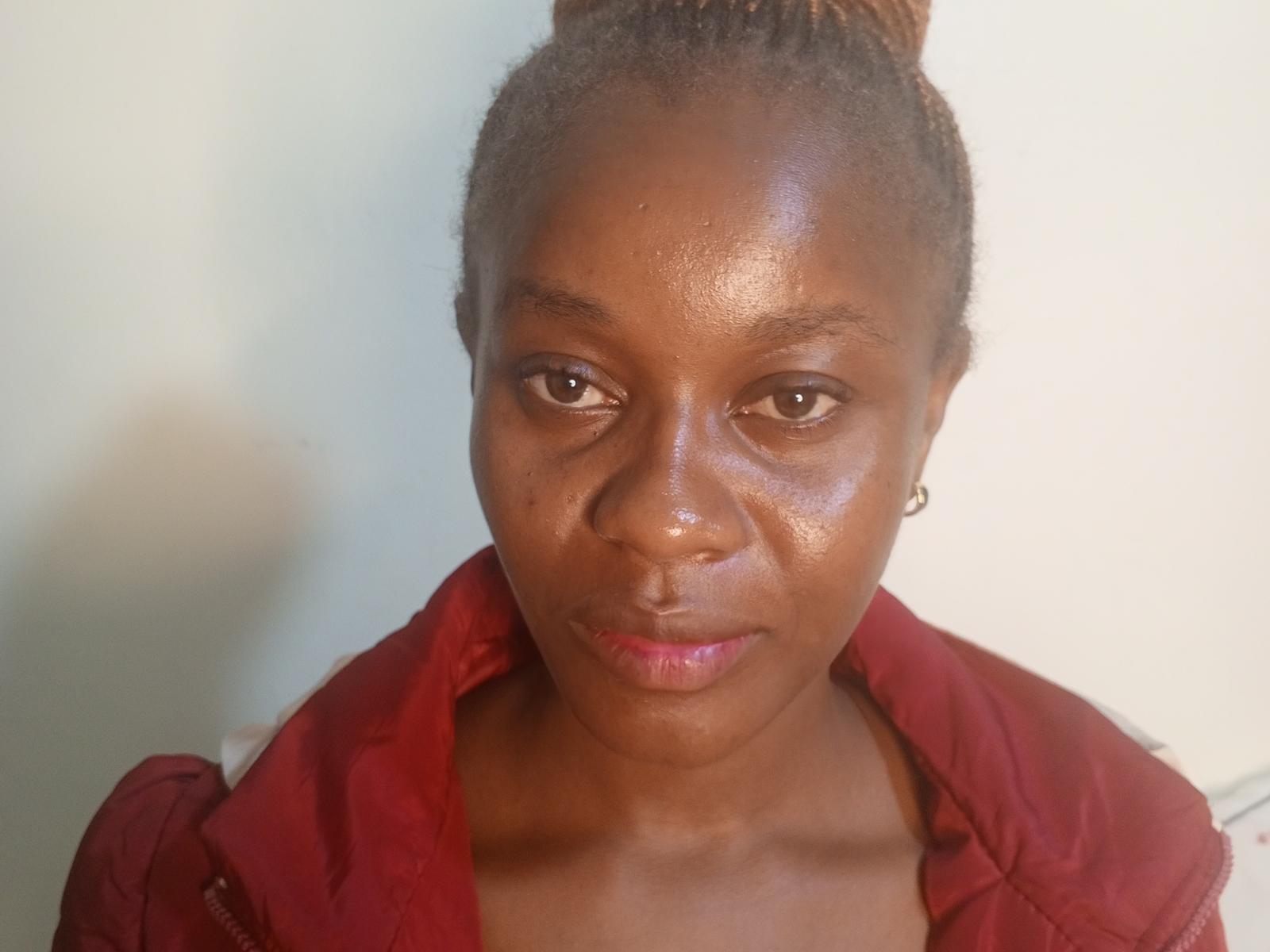 BrownieLush has dark brown skin, small earrings, a red jacket, and a slightly glossy face with a subtle frown. She's looking straight at the camera with a neutral expression.