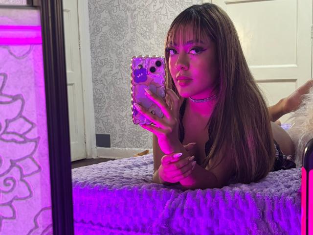 AmelieDuvall with long brown hair, lying on a purple blanket, taking a selfie with a phone in a mirror, wearing a black lace top, purple lighting.