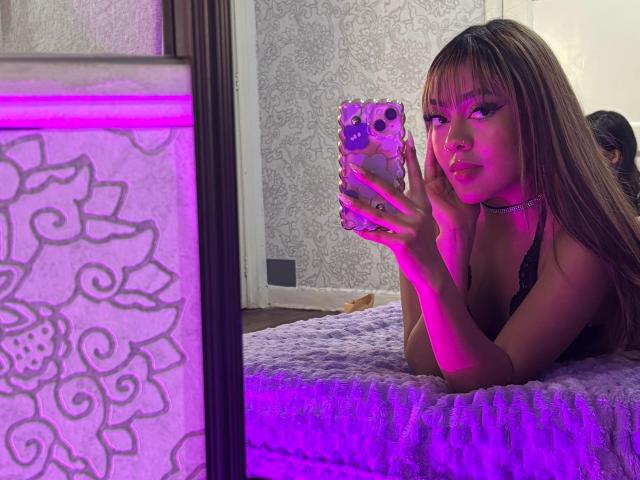 AmelieDuvall with long brown hair, black lace top, purple light, holding phone with purple case, lying on white textured bed, looking at mirror, hands on cheeks, pouty expression.
