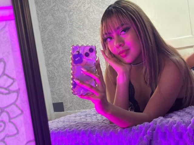 AmelieDuvall lying on a bed, holding a phone with purple case, looking in a mirror, wearing black lace top, choker, and pink eyeliner, with hands on cheek.