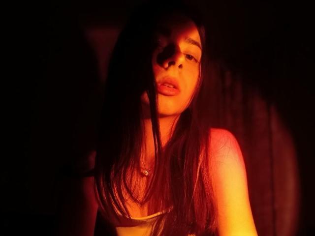 WollyMolly com cabelos longos e escuros, luz laranja brilhante, rosto parcialmente sombreado, boca ligeiramente aberta, vestindo uma blusa branca, fundo escuro, olhando para frente, luz suave no lado direito..