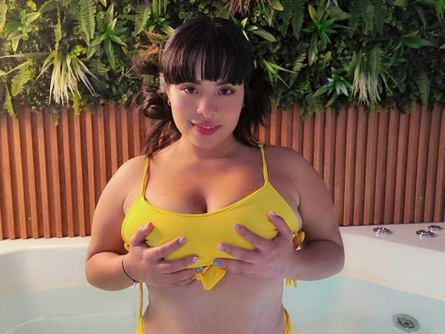 NamiOkawa with medium brown skin and dark hair, wearing a yellow bikini, holding her big tits with wet hands in a hot tub, looking at the camera.